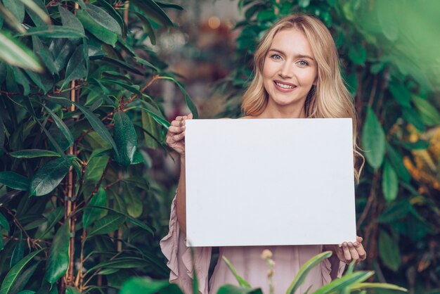 Retrato de una mujer joven rubia sonriente que se coloca en el vivero de la planta que muestra el cartel en blanco blanco