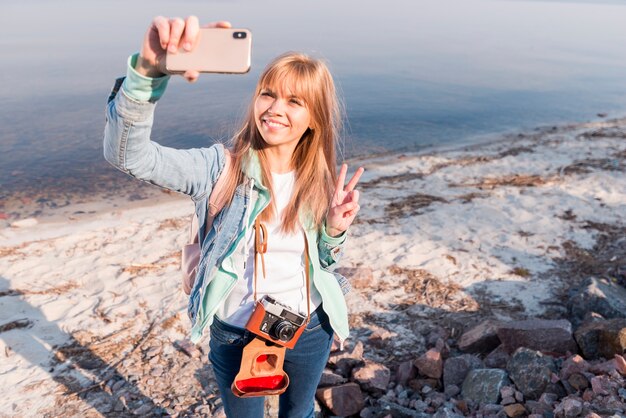 Retrato de una mujer joven rubia sonriente que hace el gesto de la paz que toma el selfie en el teléfono móvil