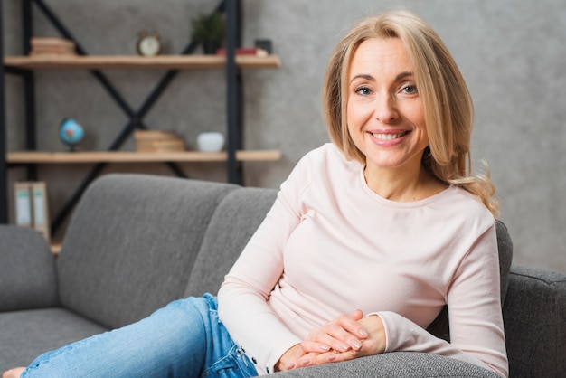 Retrato de una mujer joven rubia sonriente que se sienta en el sofá que mira la cámara