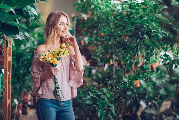 Foto gratuita retrato de una mujer joven rubia sonriente que sostiene la flor amarilla que mira lejos