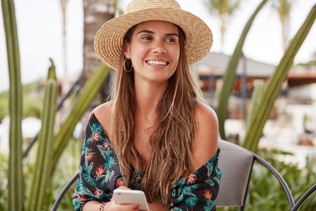 Retrato de mujer joven satisfecha con expresión positiva usa sombrero de verano y blusa elegante, se sienta en una silla cómoda al aire libre, sostiene el teléfono celular, navega por las redes sociales, descarga fotos en la página
