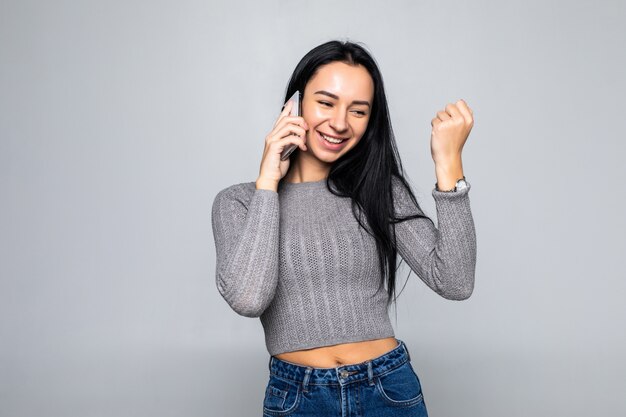 Retrato de una mujer joven satisfecha con teléfono móvil y celebrando sobre pared gris