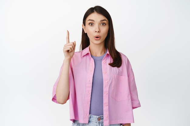 Retrato de mujer joven señalando con el dedo hacia arriba con aspecto curioso mostrando un anuncio interesante de pie sobre fondo blanco.