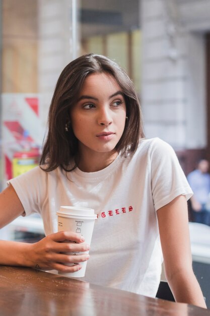 Retrato de una mujer joven sentada en una cafetería con una taza de café para llevar en la mano y mirando a otro lado