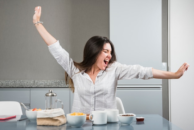 Foto gratuita retrato de una mujer joven sentada en la mesa del desayuno estirando sus brazos