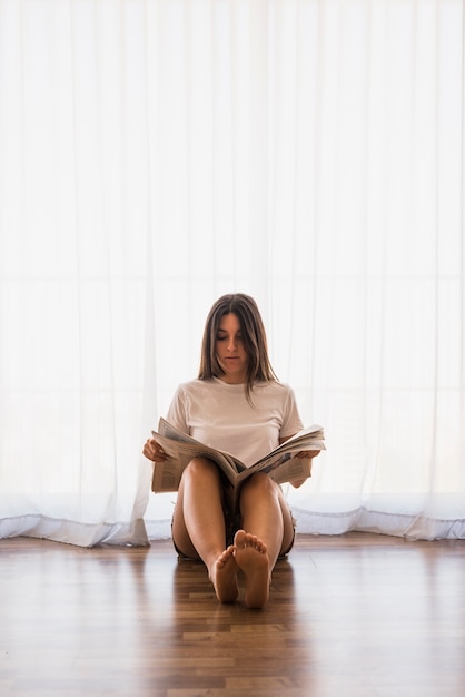 Retrato de mujer joven sentada en el piso de madera dura leyendo el periódico