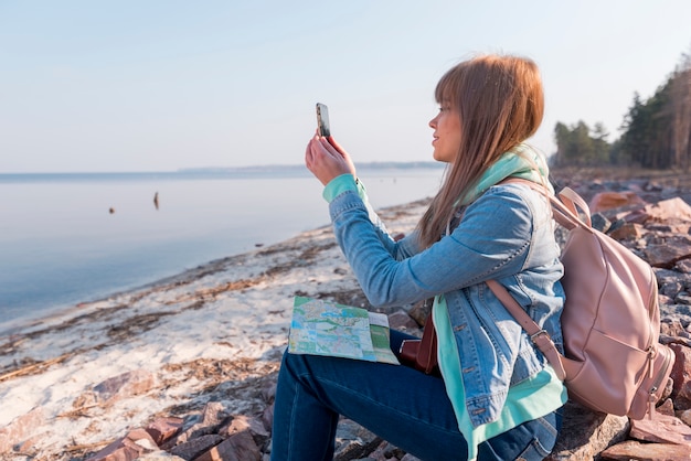 Retrato de una mujer joven sentada en la playa con el mapa usando el teléfono móvil