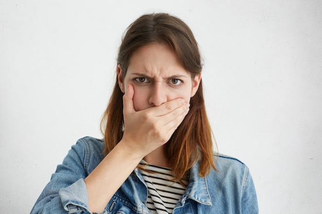 Foto gratuita retrato de mujer joven seria en chaqueta de mezclilla que cubre la boca con la mano mientras oculta el secreto