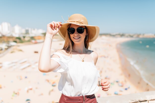Retrato de mujer joven con sombrero y gafas de sol redondas, clima ventoso agradable día de verano en el océano