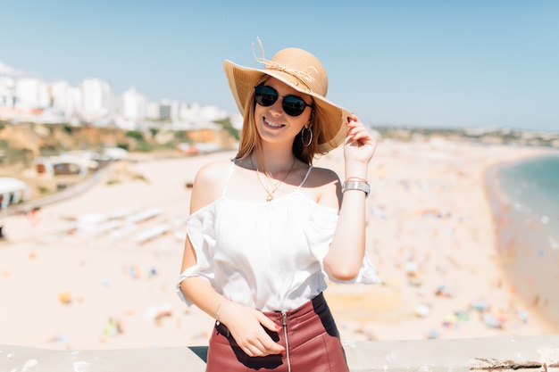 Retrato de mujer joven con sombrero y gafas de sol redondas, clima ventoso agradable día de verano en el océano