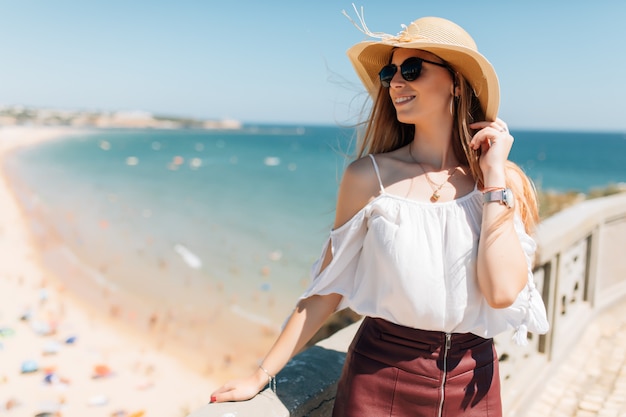 Retrato de mujer joven con sombrero y gafas de sol redondas, clima ventoso agradable día de verano en el océano