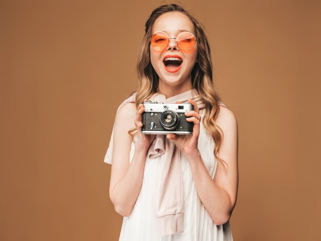 Retrato de la mujer joven sonriente alegre que toma la foto con la inspiración y que lleva el vestido blanco. Chica sosteniendo la cámara retro. Modelo en gafas de sol posando