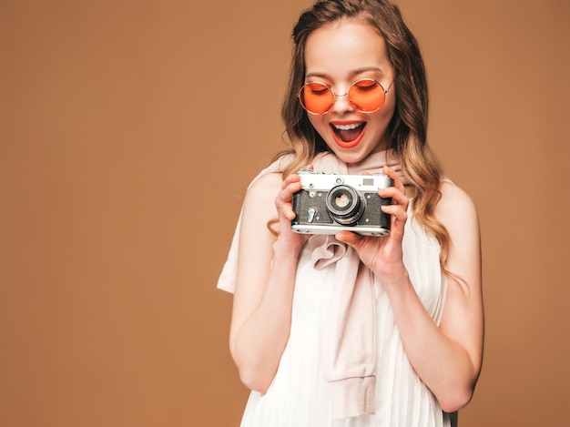 Retrato de la mujer joven sonriente alegre que toma la foto con la inspiración y que lleva el vestido blanco. Chica sosteniendo la cámara retro. Modelo en gafas de sol posando