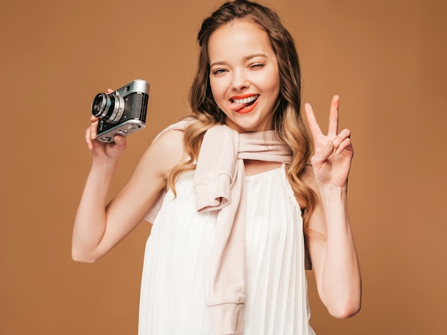 Retrato de la mujer joven sonriente alegre que toma la foto con la inspiración y que lleva el vestido blanco. Chica sosteniendo la cámara retro. Modelo posando, mostrando el signo de paz