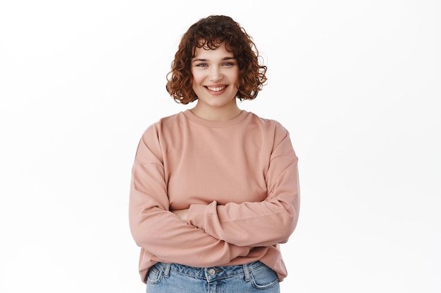 Retrato de mujer joven sonriente confiada, brazos cruzados en el pecho, sonrisas y miradas con determinación y expresión segura de sí misma, de pie sobre fondo blanco.