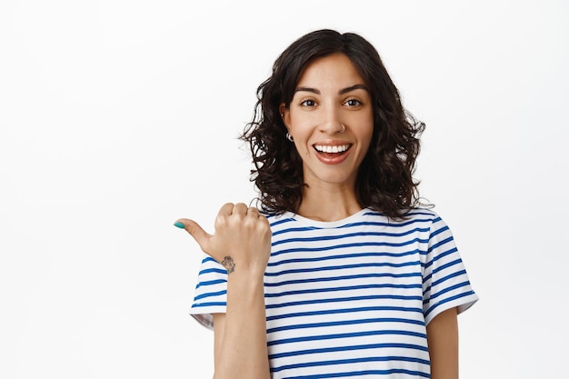 Retrato de mujer joven sonriente con dientes blancos, nariz perforada, apuntando a la izquierda en la pancarta y mirando feliz a la cámara, mostrando texto de promoción sobre fondo blanco vacío