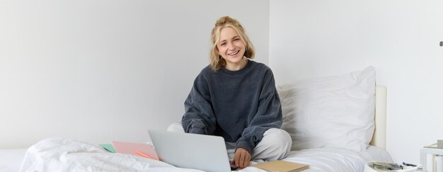 Retrato de una mujer joven sonriente estudiando en su cama trabajando desde casa en el dormitorio sentada con una computadora portátil