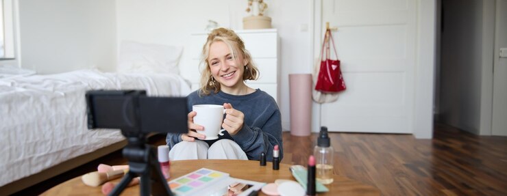 Foto gratuito retrato de una mujer joven sonriente grabando video en la cámara sosteniendo una taza de té hablando haciendo