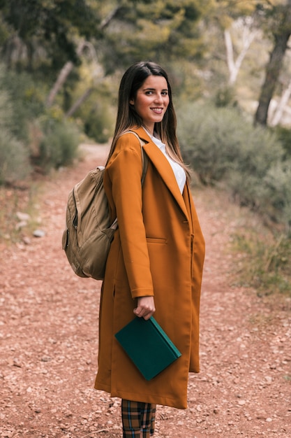 Foto gratuita retrato de una mujer joven sonriente con mochila sosteniendo el libro en la mano