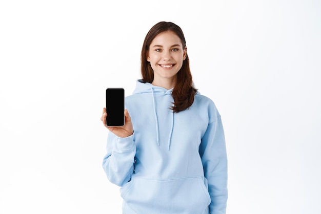 Retrato de una mujer joven sonriente mostrando teléfono móvil de pantalla en blanco aislado sobre fondo blanco.