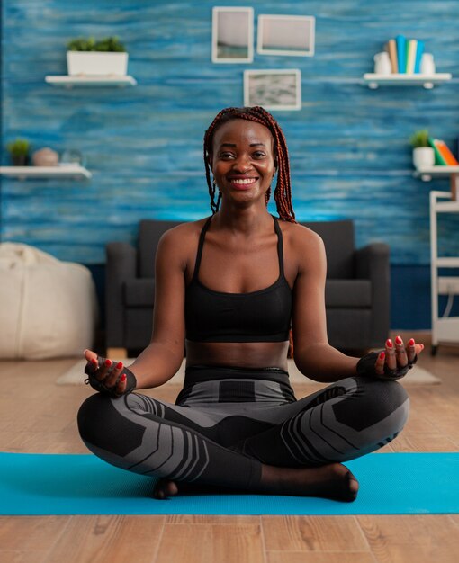 Retrato de mujer joven sonriente practicando yoga en casa sentado en posición de loto en la estera meditando, practicando la atención plena