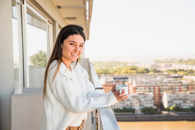 Retrato de una mujer joven sonriente que se coloca en el balcón que sostiene la taza del café con leche