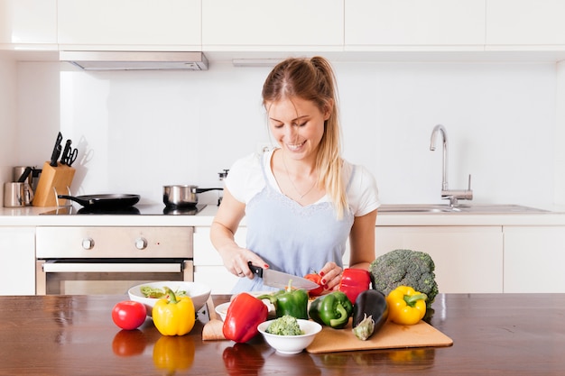 Retrato de una mujer joven sonriente que corta las verduras frescas con el cuchillo en la tabla de madera