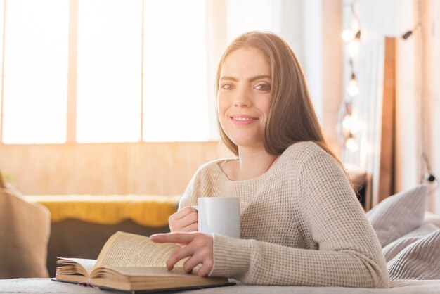 Retrato de una mujer joven sonriente que mira a la cámara mientras que lee el libro en casa