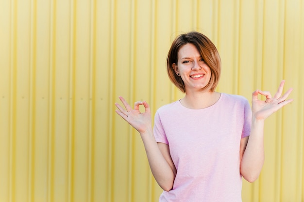 Foto gratuita retrato de una mujer joven sonriente que muestra gesto de mano contra fondo amarillo del obturador