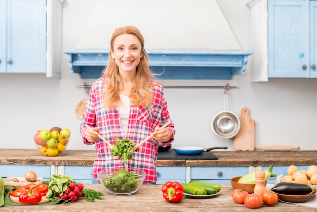 Retrato de una mujer joven sonriente que prepara la ensalada de verduras en la cocina
