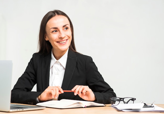 Retrato de la mujer joven sonriente que se sienta en el lugar de trabajo que mira lejos