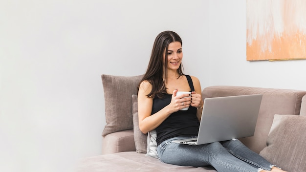 Foto gratuita retrato de una mujer joven sonriente que se sienta en el sofá que sostiene la taza de café en la mano que mira el ordenador portátil