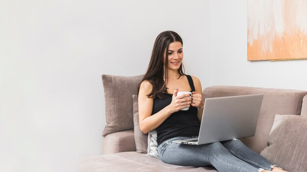 Retrato de una mujer joven sonriente que se sienta en el sofá que sostiene la taza de café en la mano que mira el ordenador portátil