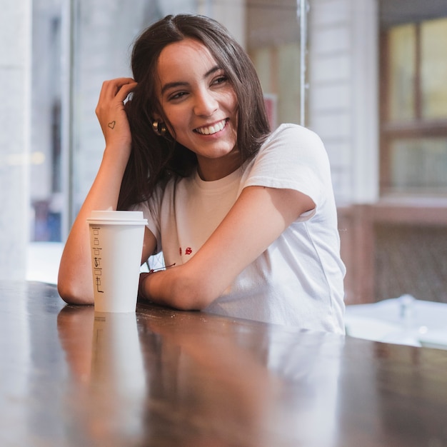 Retrato de una mujer joven sonriente que se sienta en la tabla con la taza de café para llevar en la tabla de madera