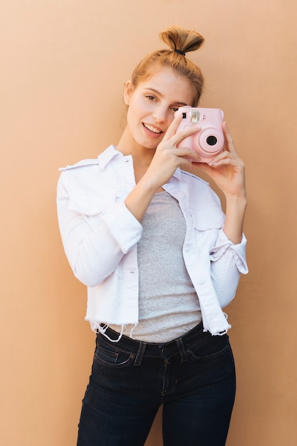 Retrato de una mujer joven sonriente que sostiene la cámara instantánea rosada contra el contexto beige