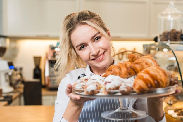Retrato de una mujer joven sonriente que sostiene el croissant cocido en el soporte de la torta