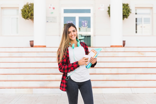 Retrato de una mujer joven sonriente que sostiene los libros en la mano que se opone al edificio de la universidad