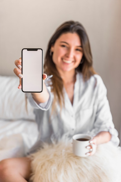 Retrato de una mujer joven sonriente que sostiene la taza de café que muestra la pantalla del teléfono inteligente