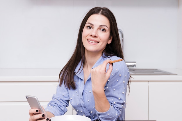 Retrato de una mujer joven sonriente que usa el teléfono móvil que sostiene la galleta en la mano