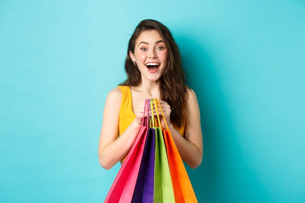 Retrato de mujer joven sorprendida mira asombrada a la cámara, sosteniendo bolsas de la compra, ver descuentos en la tienda, de pie sobre fondo azul.