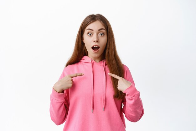 Retrato de mujer joven sorprendida mira con incredulidad, boca abierta jadeando al frente, apuntando a sí misma, ganando un premio o siendo elegida, pared blanca