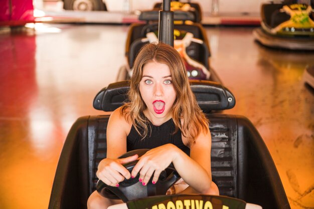 Retrato de la mujer joven sorprendida que se sienta en el coche de parachoques en el parque de atracciones