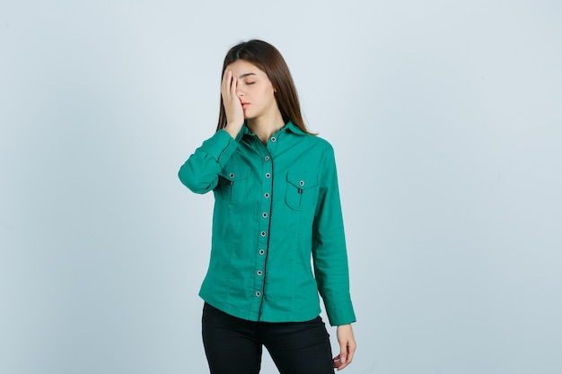 Retrato de mujer joven sosteniendo la mano en la cara con camisa verde, pantalones y mirando cansado vista frontal