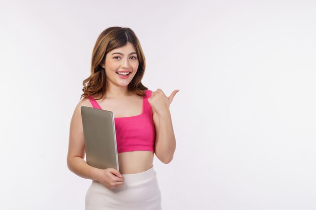 Retrato de mujer joven sosteniendo un ordenador portátil y señalando con el dedo a un lado aislado sobre fondo blanco.