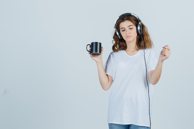 Retrato de mujer joven sosteniendo una taza de bebida en camiseta blanca y mirando la vista frontal de ensueño