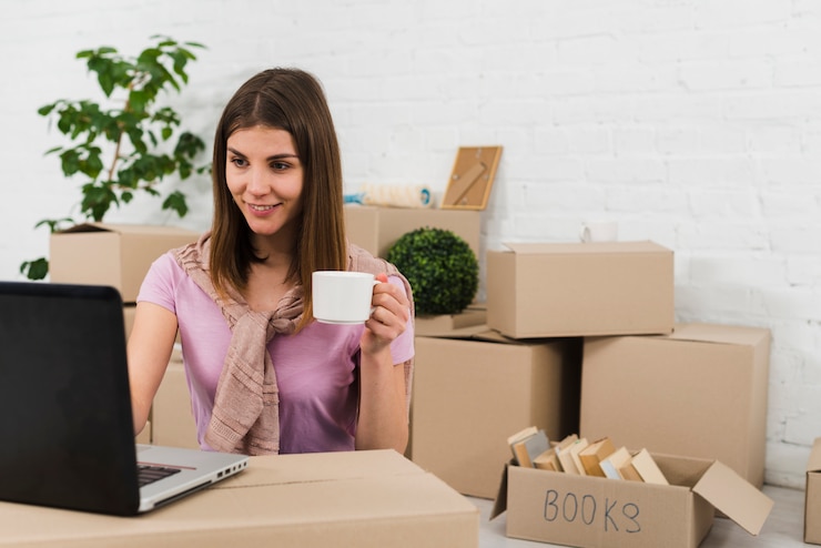 Foto gratuito retrato de una mujer joven sosteniendo una taza de café en la mano usando una computadora portátil en su nueva casa
