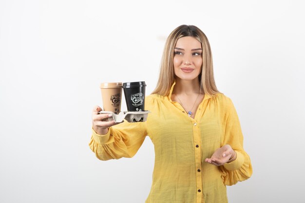 Retrato de mujer joven sosteniendo tazas de café y de pie sobre una pared blanca.