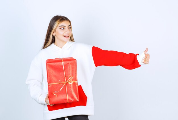 Retrato de mujer joven en suéter caliente con regalo de Navidad mostrando el pulgar hacia arriba
