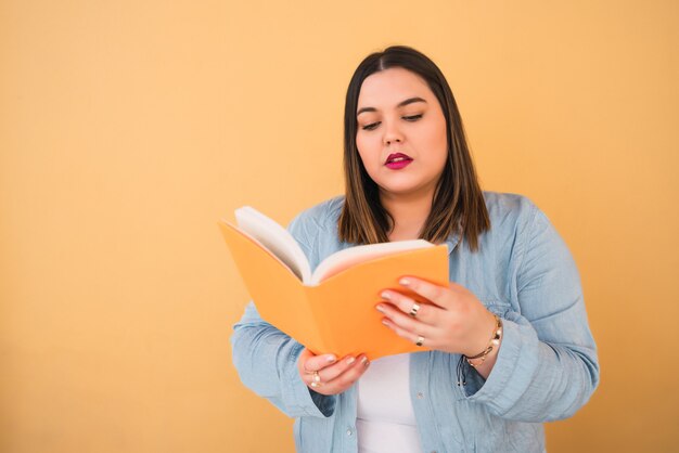 Retrato de mujer joven de talla grande disfrutando de tiempo libre y leyendo un libro mientras está de pie contra el espacio amarillo. Concepto de estilo de vida.