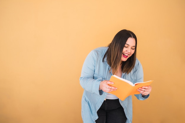 Foto gratuita retrato de mujer joven de talla grande disfrutando de tiempo libre y leyendo un libro mientras está de pie contra el fondo amarillo. concepto de estilo de vida.